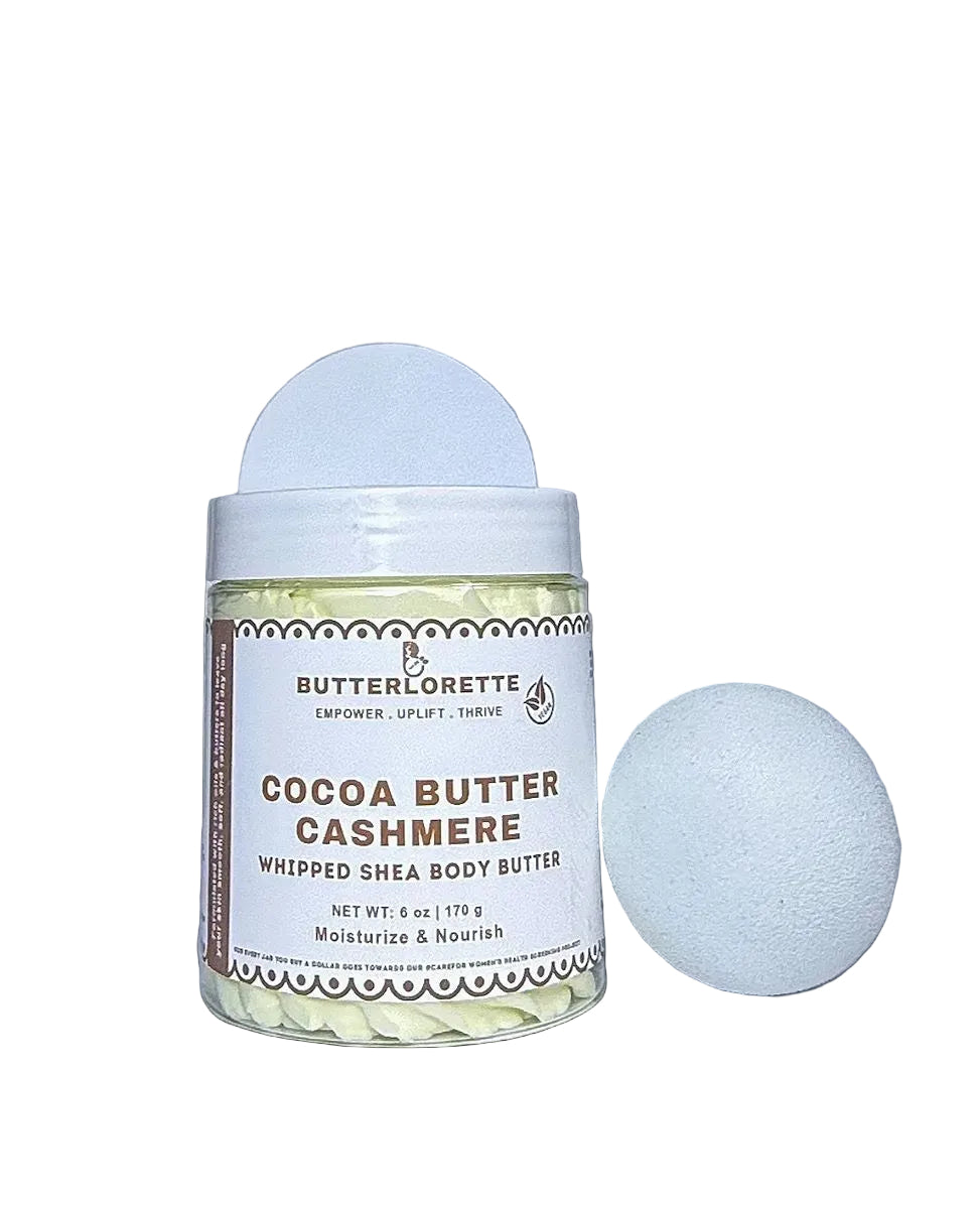 A jar of ButterLorette Cocoa Butter Cashmere whipped Shea body butter next to a white bath bomb sponge on a light gray background.
