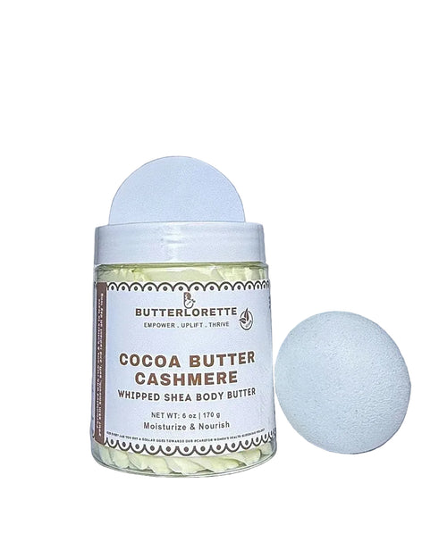 A jar of ButterLorette Cocoa Butter Cashmere whipped Shea body butter next to a white bath bomb sponge on a light gray background.